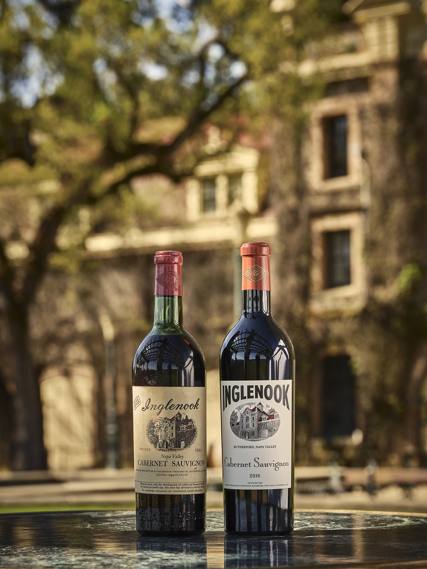 Two bottles, one old and one current, sitting on a green marble table. Behind the bottles is the stone Chateau and a large oak tree. 